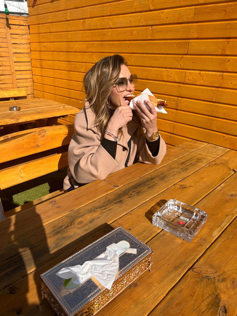 Žena jede sendvič za drvenim stolom na otvorenom